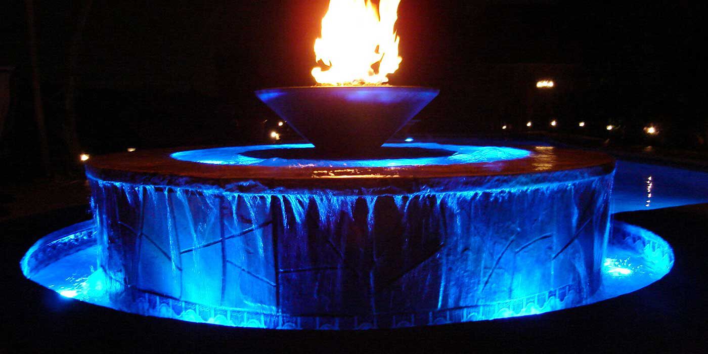 A large outdoor water fountain at night with vibrant blue LED lighting and a large fire bowl installed on top.