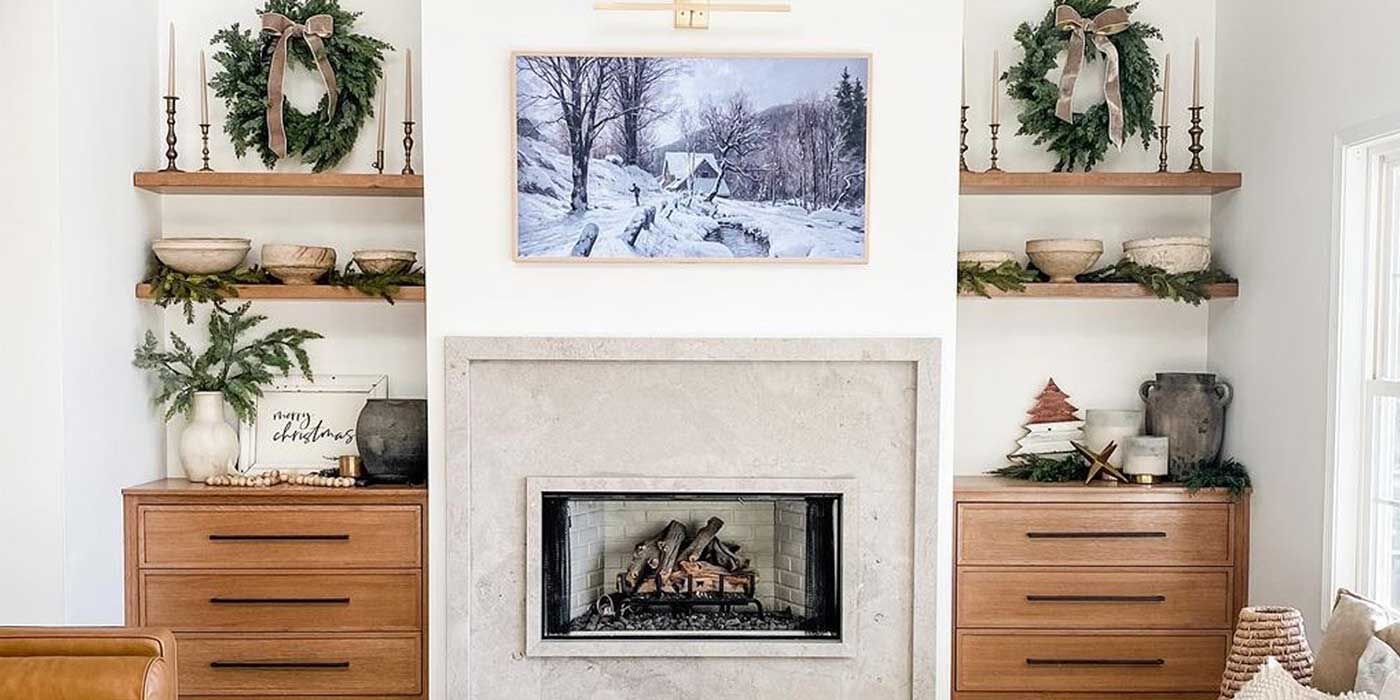 A modern farmhouse-inspired fireplace mantel with built-in wooden shelves and drawers decorated with organic greenery and simple wooden accent pieces, a large painting of a winter landscape, and a rectangular wood burning fireplace.