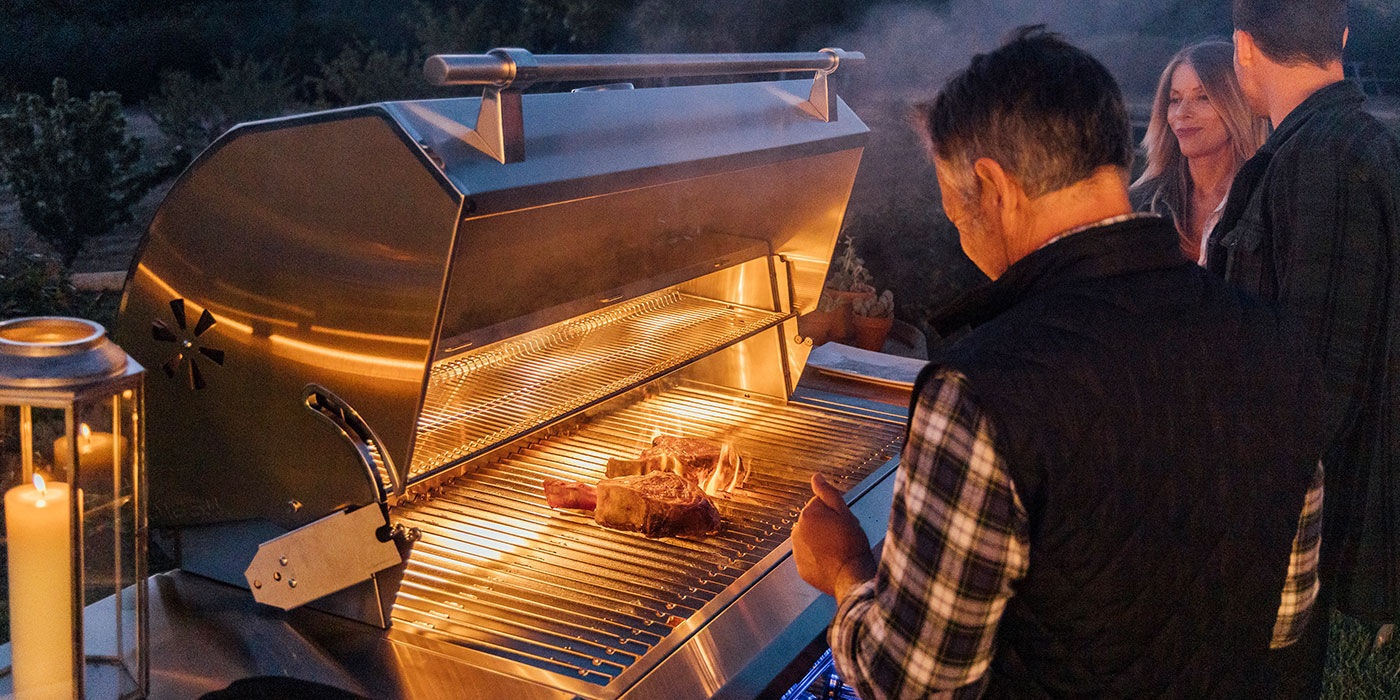 A group of people standing in front of an open grill with interior halogen lights at night, cooking steaks.