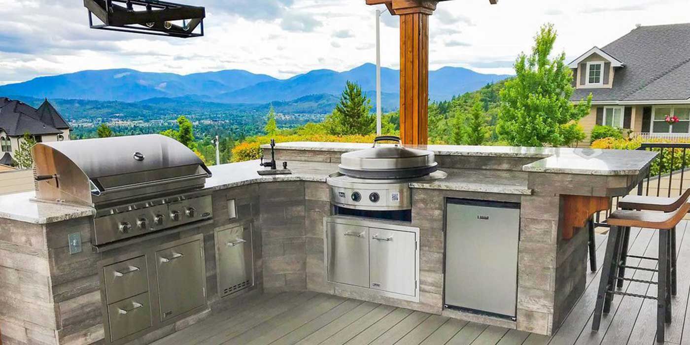 A large, custom outdoor kitchen island with a built-in grill, kamado grill, built in stainless steel storage doors and drawers, and an outdoor refrigerator, sink, and faucet.
