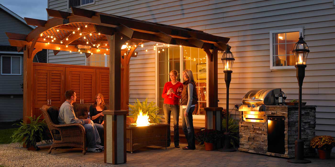 A group of people gathered on an outdoor patio at night with a wooden gazebo, a gas fire pit, an outdoor kitchen island and grill, and tiki torches.