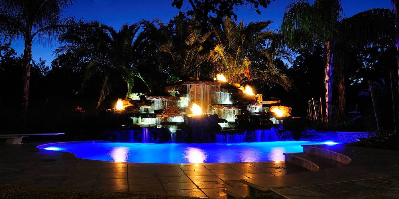 A private island grotto at night with layered rock walls, lush greenery, vibrant blue LED accent lighting, and several fire and water bowls installed around a pool.