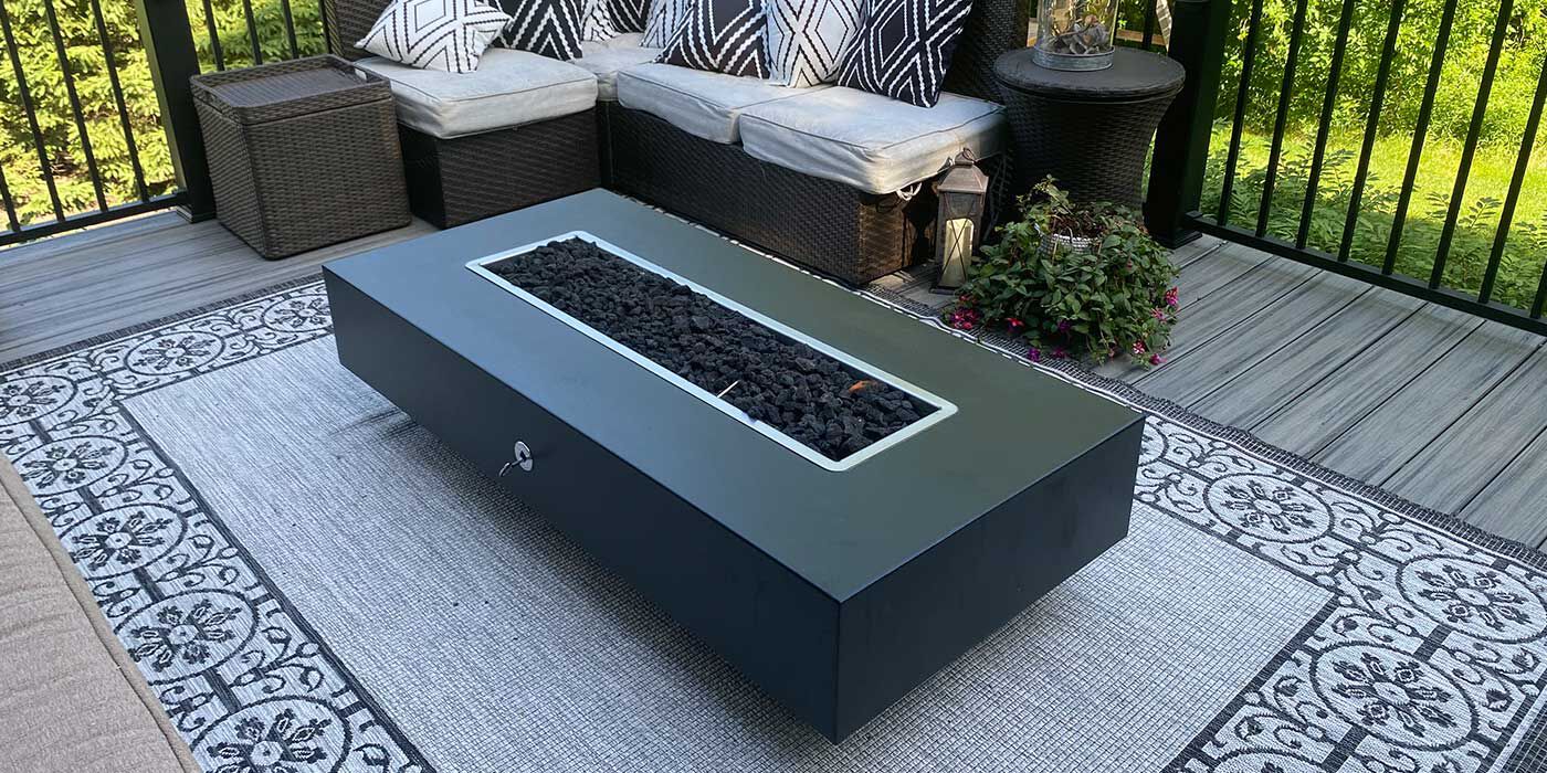 A large, linear black gas fire pit placed on a wooden balcony with a large sectional couch nearby