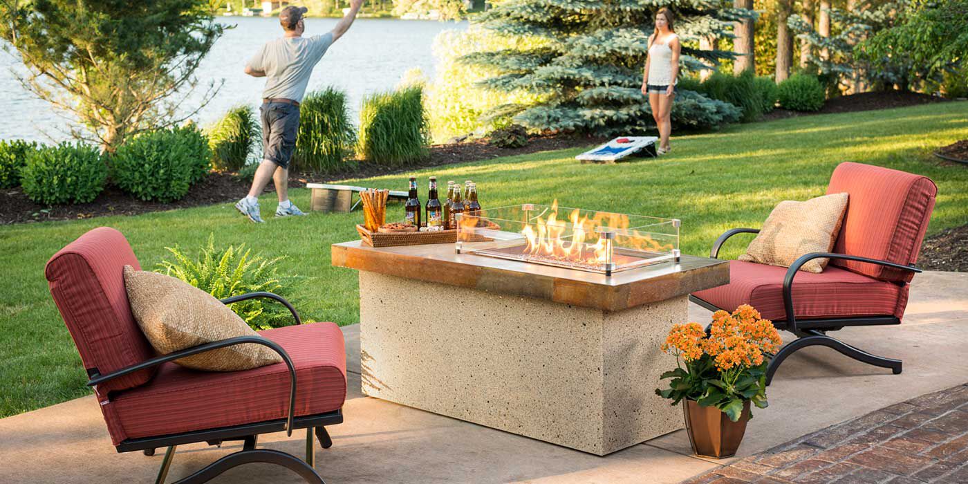 An inviting backyard scene showcases a cozy outdoor seating area centered around a modern propane fire pit table. The fire pit features a large, rectangular stone base with a metal top and glass wind guards, from which flames elegantly rise. 