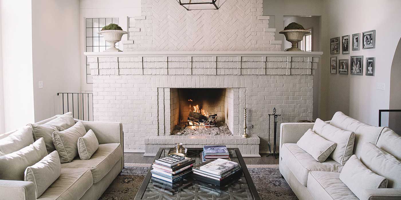A transitional living room with white-painted, exposed brick walls, white plush couches, and a large hearth with a classic, square fireplace.