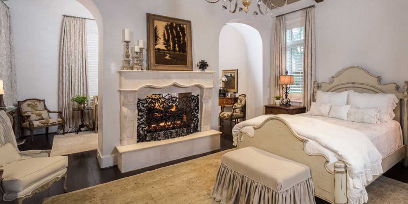 A French country-inspired bedroom with a connected sitting area, a large beige and white bed with a decorative bedframe, and a large white hearth with a traditional, wood burning fireplace.