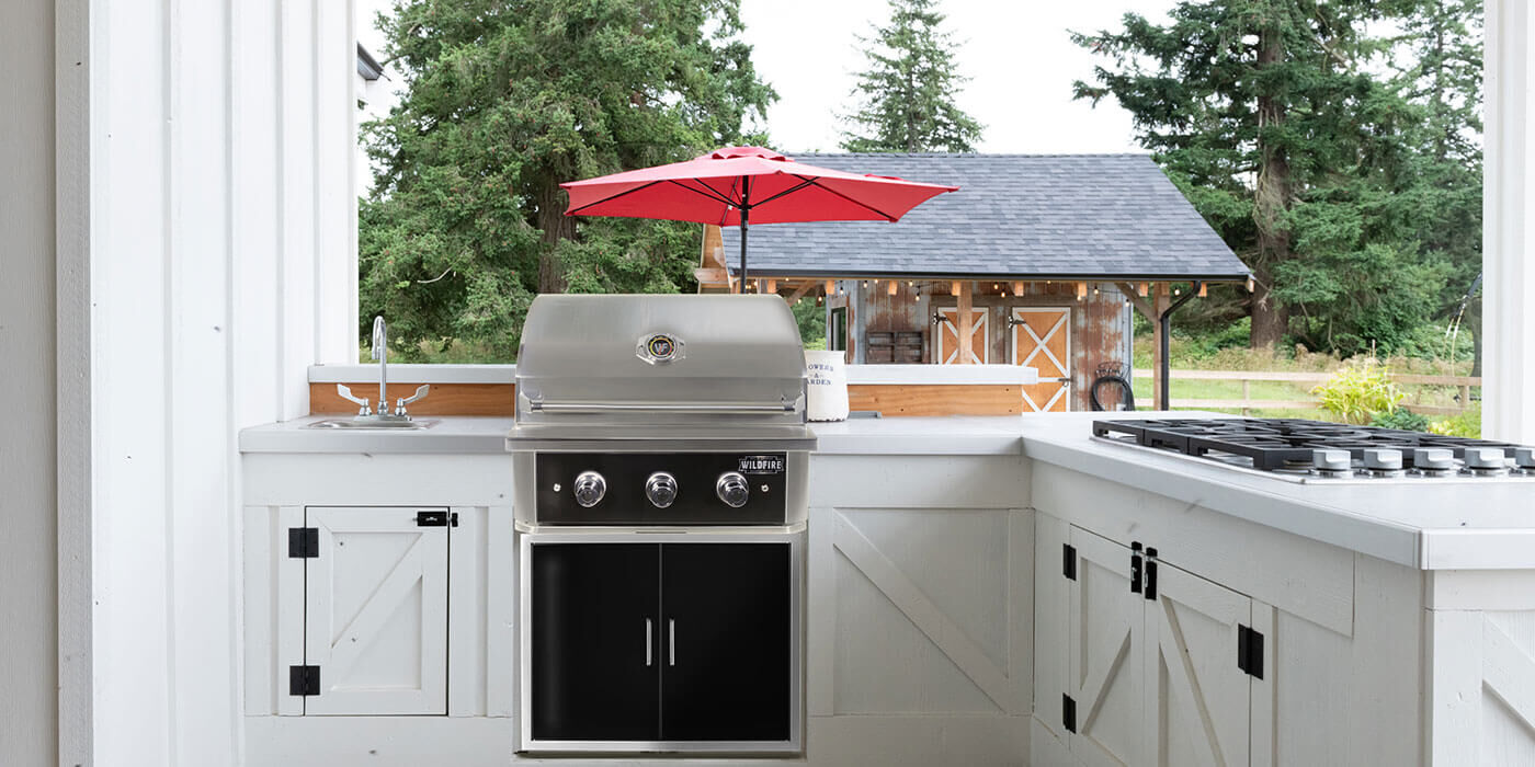 An outdoor kitchen island with white, farmhouse-style cabinets with a crisscross pattern, and a built-in grill with black stainless steel storage doors.