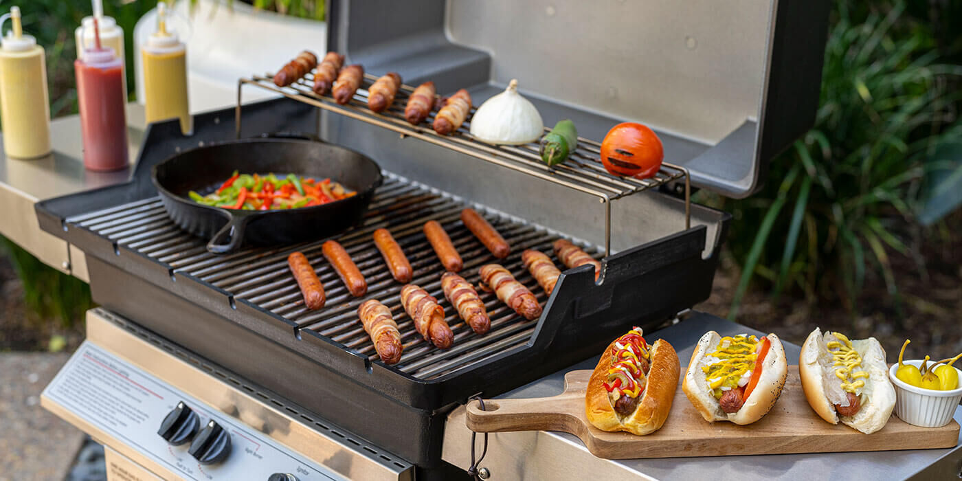 A close-up view of a small gas post-mounted grill cooking hot dogs and warming up veggies.