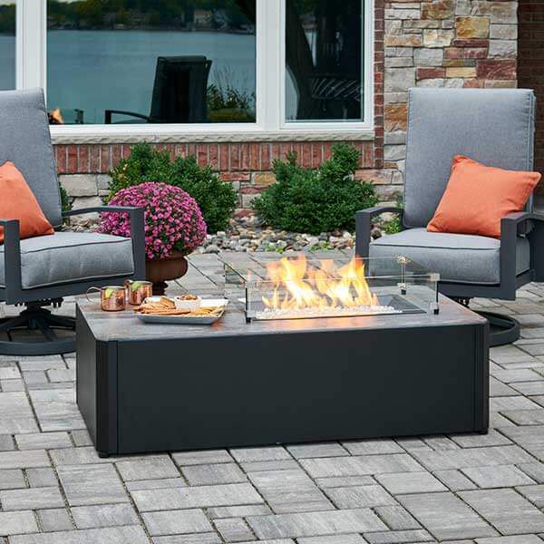 A black fire pit table and two chairs sitting on top of a stone paved patio.