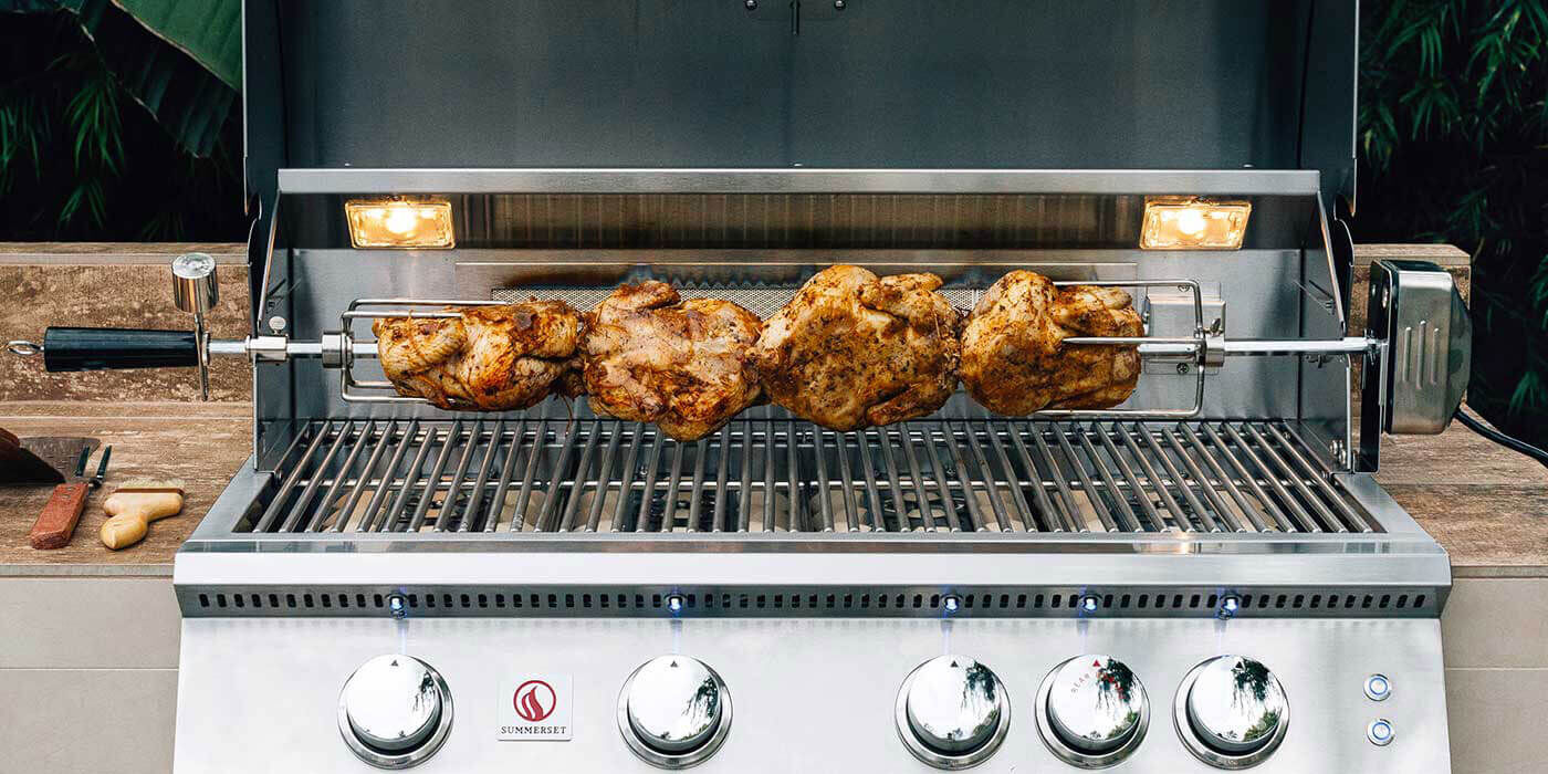 A stainless steel gas grill with a motorized rotisserie kit cooking four small whole chickens.