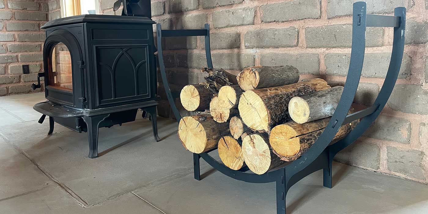 A stack of seasoned firewood on a small crescent shaped firewood rack next to a black wood stove.