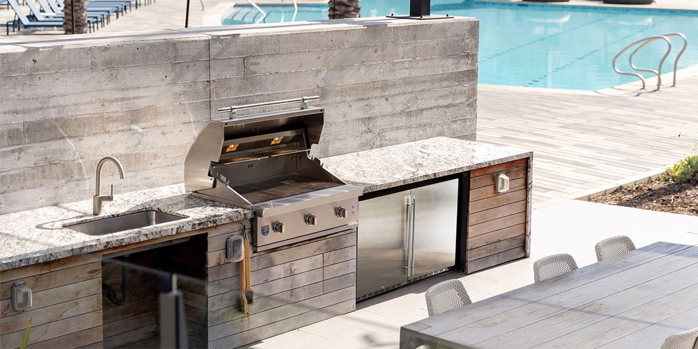 An outdoor kitchen island with a built-in stainless steel gas grill and an outdoor refrigerator installed on a patio near a pool.