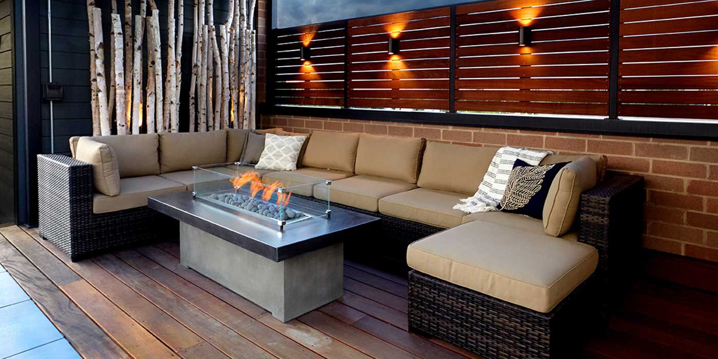 A contemporary outdoor space with a wooden deck, a large sectional couch, and a linear gas fire pit with a glass wind guard