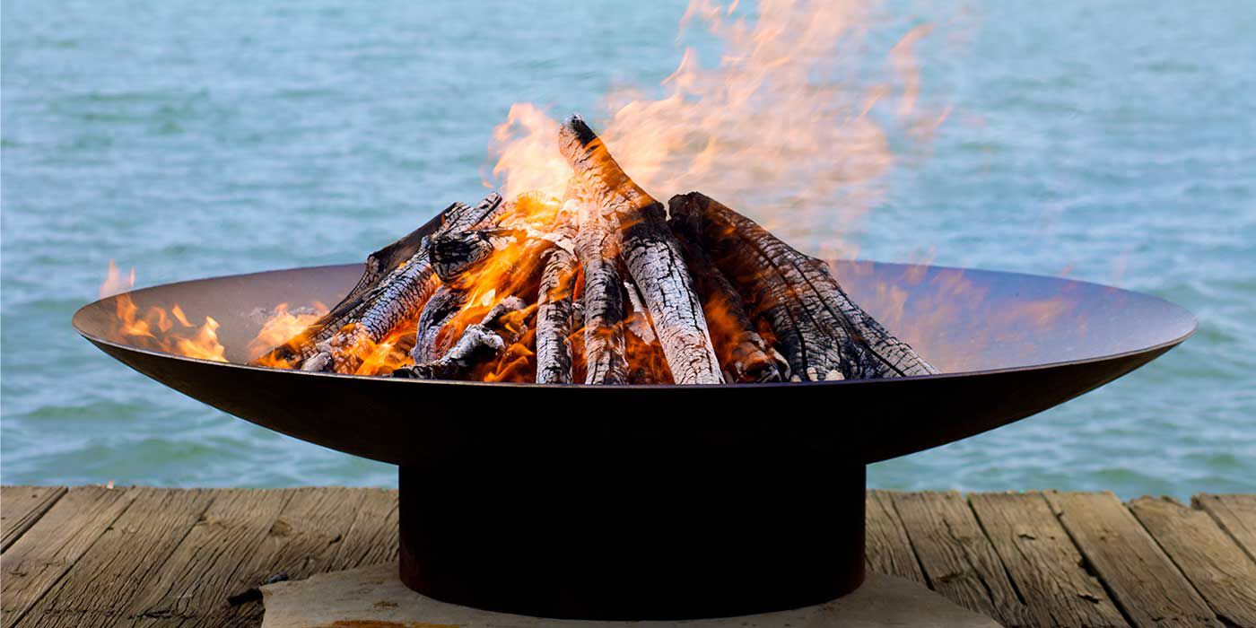 A large fire bowl with burning logs placed on a wooden deck overlooking a body of water.