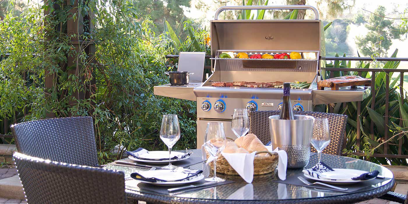 An outdoor patio with a dining table and chairs, and a large, cart-mount gas grill cooking various meats and vegetables.
