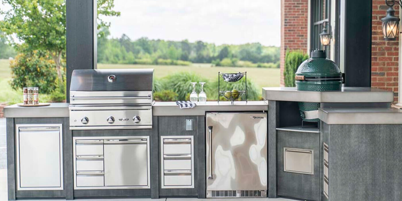 A large, custom outdoor kitchen island with stainless steel storage components, a built-in, stainless steel grill head, an outdoor refrigerator and sink, and a green kamado grill installed into the countertop.