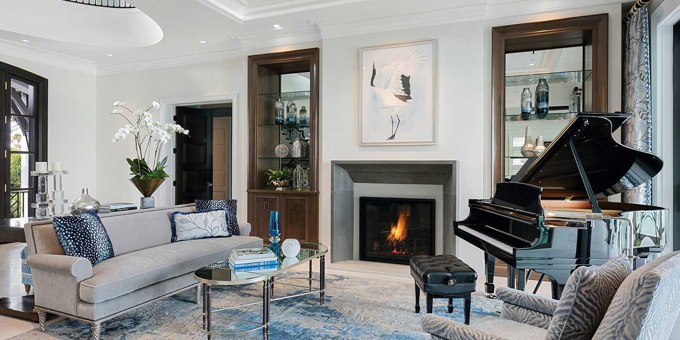 A traditional living room with white walls, dark wood trim, a grey couch and accent chair, dark blue accent pillows, a blue and gray area rug, a large black piano, and a square gas fireplace with a black and white line drawing hanging over it.