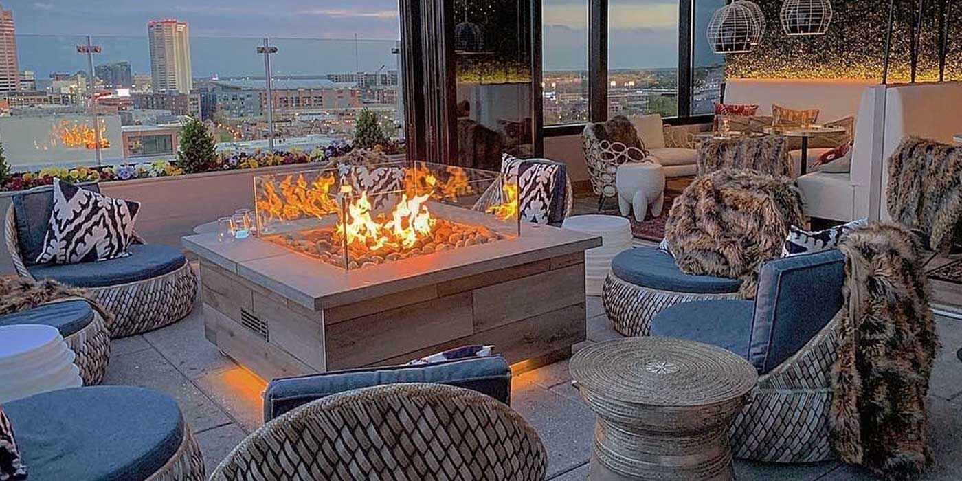 A modern bohemian outdoor lounging space on the rooftop in the city with a large, square gas fire pit and eight wicker chairs with faux fur blankets, blue cushions, and geometric-patterned pillows.