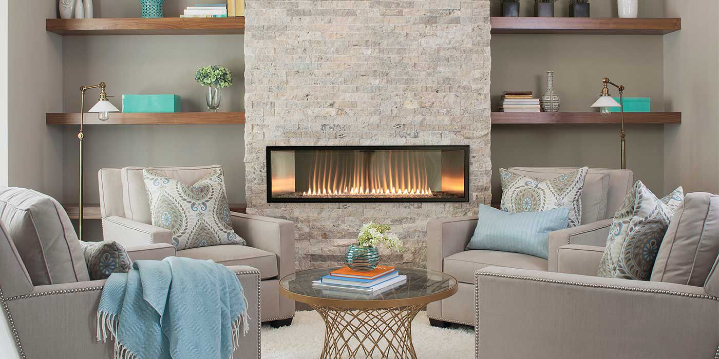 A transitional living room with beige walls, built-in wooden shelves, a large natural stone hearth with a linear Empire gas fireplace, four light gray chairs, and a round, bronze coffee table.