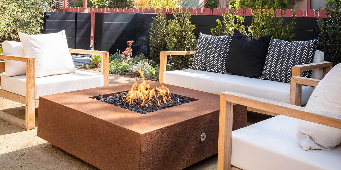 A cozy outdoor seating area features a modern natural gas fire pit at the center, surrounded by light wooden frame armchairs and a matching loveseat with white cushions. 